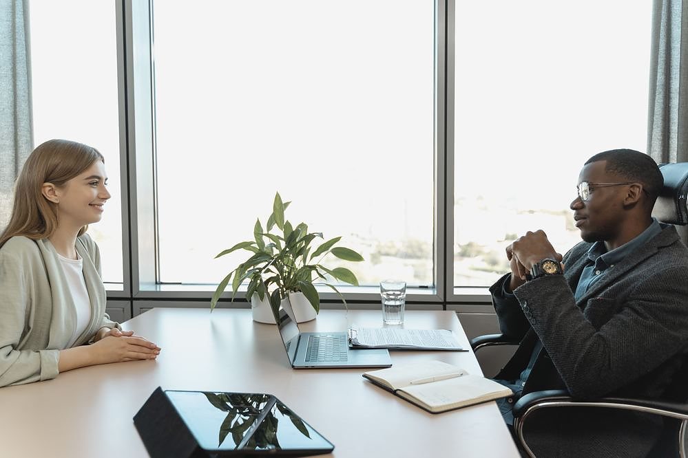 Two people sitting across a table in a daylit meeting room, mid-conversation